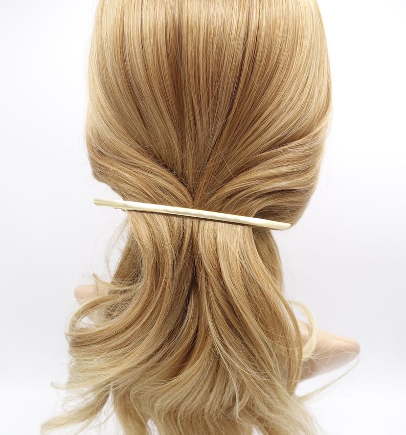 May include: A long, slender, gold-toned metal hair barrette with a smooth, slightly curved finish. The hair accessory is positioned horizontally across the back of a head of light blonde hair, securing a section of the hair in place.