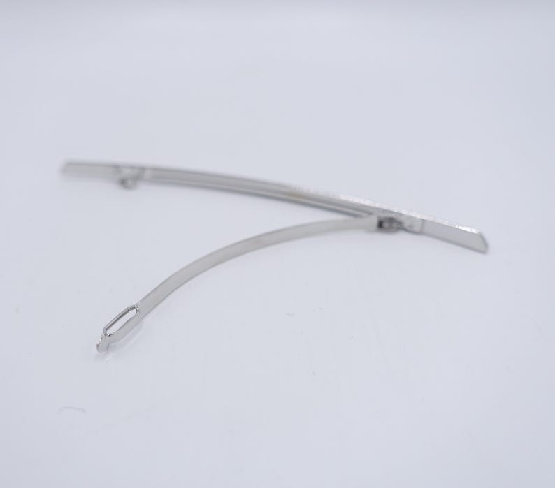 May include: A silver-toned metal hair barrette featuring a long, slightly curved base bar and a thinner, flexible hinged clasp arm that extends from one side. The mechanism includes a small loop at the end of the clasp for securing the hair accessory.