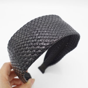 Pode incluir: Uma faixa de cabeça preta tecida com um design largo e plano. A faixa de cabeça é feita de um material texturizado que se assemelha ao couro.