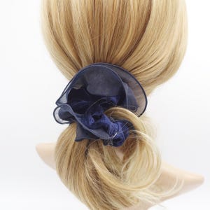 May include: A navy blue scrunchie with a layered, ruffled design. The scrunchie is made of a sheer fabric and a velvet-like material, creating a textured and elegant look. The scrunchie is wrapped around a blonde ponytail.
