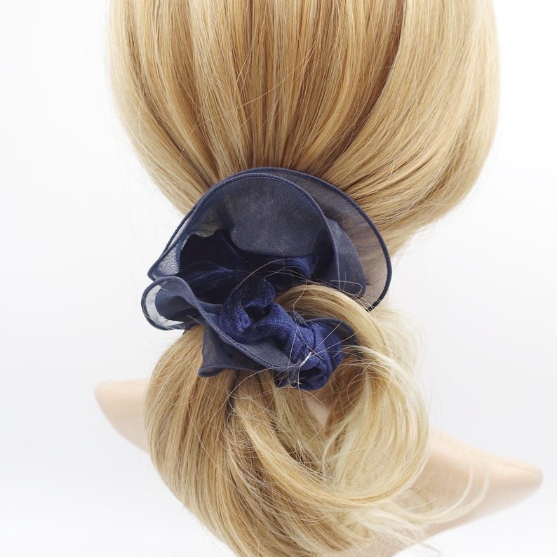 May include: A navy blue scrunchie with a layered, ruffled design. The scrunchie is made of a sheer fabric and a velvet-like material, creating a textured and elegant look. The scrunchie is wrapped around a blonde ponytail.