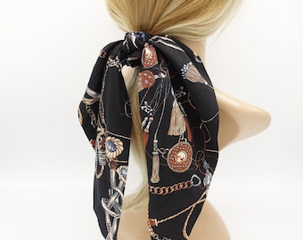 May include: Black hair tie with a patterned scarf featuring gold chains, tassels, and ornate designs. The scarf is tied in a bow at the back of the head.