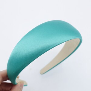 May include: A teal satin headband with a padded, rounded top and a light beige interior. The headband is a solid color and appears to be made of a smooth, shiny fabric. The headband is a fashion accessory.