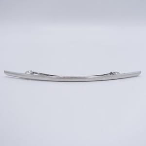 May include: A long, slender, silver-toned metal hair barrette with a smooth, slightly curved design. The accessory features a functional spring-loaded clasp mechanism on the back, designed to secure hair in place with a polished, metallic finish.