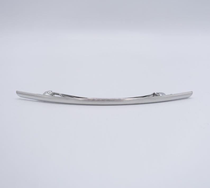 May include: A long, slender, silver-toned metal hair barrette with a smooth, slightly curved design. The accessory features a functional spring-loaded clasp mechanism on the back, designed to secure hair in place with a polished, metallic finish.