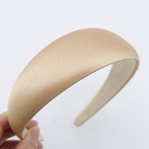 May include: A beige satin headband with a padded design. The headband is a neutral color and has a smooth, shiny texture. The interior is a lighter beige color. The headband is held by a hand against a white background.