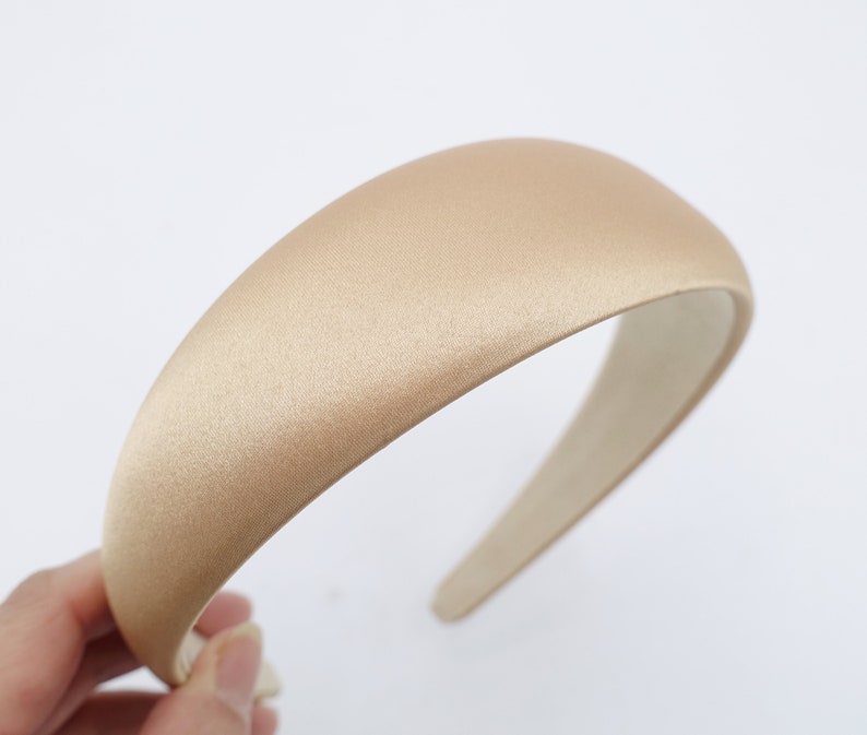 May include: A beige satin headband with a padded design. The headband is a neutral color and has a smooth, shiny texture. The interior is a lighter beige color. The headband is held by a hand against a white background.