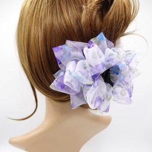 Puede incluir: Una pinza para el pelo floral blanca y morada con un gran diseño de volantes. La pinza está hecha de tela transparente y tiene un aspecto delicado y femenino.