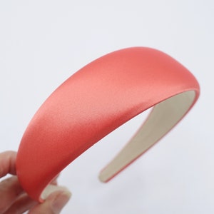 May include: A coral-colored satin headband with a padded, rounded top. The headband has a smooth, lustrous finish and a neutral-toned interior. This hair accessory is designed for comfort and style.