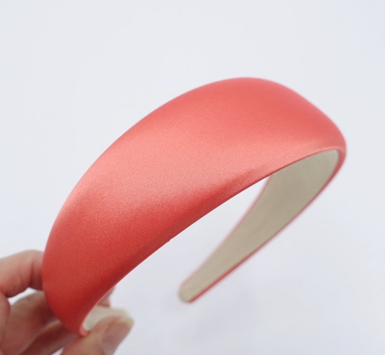 May include: A coral-colored satin headband with a padded, rounded top. The headband has a smooth, lustrous finish and a neutral-toned interior. This hair accessory is designed for comfort and style.