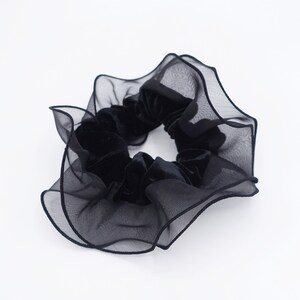 May include: A black scrunchie with a velvet center and a sheer, ruffled outer layer. The scrunchie is a hair accessory, perfect for adding a touch of elegance to any hairstyle. The fabric appears soft and delicate.