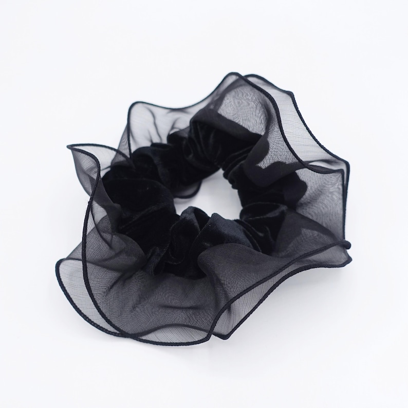 May include: A black scrunchie with a velvet center and a sheer, ruffled outer layer. The scrunchie is a hair accessory, perfect for adding a touch of elegance to any hairstyle. The fabric appears soft and delicate.