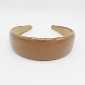 May include: A brown, padded headband with a glossy, crocodile-skin texture. The headband is a solid color and has a neutral, light brown interior. The accessory is designed to be worn on the head.
