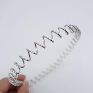 May include: A silver metal headband featuring a zig-zag pattern. The headband is crafted from a continuous wire, creating a spiral form. The design is minimalist and contemporary, suitable for different hairstyles.