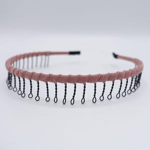 May include: A dusty rose headband with black fringe detailing. The headband features a twisted fabric design and small black fringe hanging from the bottom. The headband is a simple, yet elegant hair accessory.