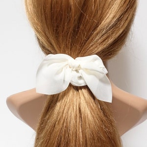 Puede incluir: Una goma de pelo de tela blanca crema con un lazo atado alrededor de una cola de caballo. La goma para el pelo está hecha de un material suave y fluido. El cabello es de color marrón claro.