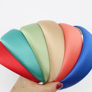 May include: A collection of padded headbands in various colors, including red, teal, green, beige, coral, and blue. The headbands are made of a smooth, satin-like material and are held in a hand against a white background. These headbands are a fashion accessory.