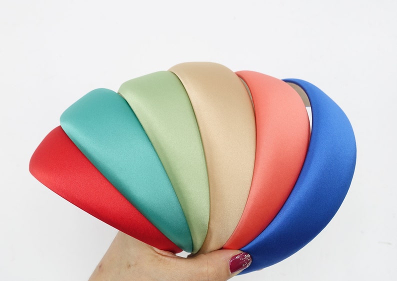 May include: A collection of padded headbands in various colors, including red, teal, green, beige, coral, and blue. The headbands are made of a smooth, satin-like material and are held in a hand against a white background. These headbands are a fashion accessory.