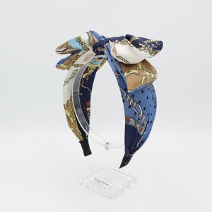 Könnte beinhalten: Ein gemustertes Stirnband mit einer großen Schleife. Das Stirnband hat eine Mischung aus blauen, cremefarbenen und braunen Farben mit Ketten- und Tupfenmustern. Die Schleife ist aus seidigem Material.