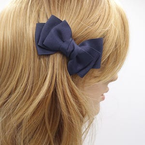 May include: A navy blue bow hair clip is attached to blonde hair. The bow is made of a ribbed fabric and is positioned on the side of the head. The hair clip is a simple yet elegant accessory.