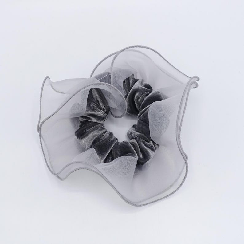 May include: A gray velvet scrunchie with a sheer, ruffled overlay. The scrunchie is a soft gray color and is surrounded by a delicate, light gray fabric. The overall design is elegant and feminine.