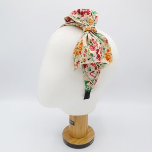 May include: A floral headband with a large bow. The headband is beige with a floral pattern in shades of red, yellow, and green. The bow is tied in a knot at the top of the headband.
