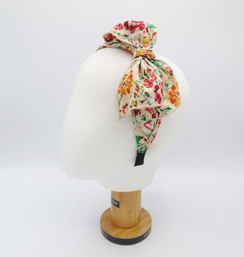 May include: A floral headband with a large bow. The headband is beige with a floral pattern in shades of red, yellow, and green. The bow is tied in a knot at the top of the headband.