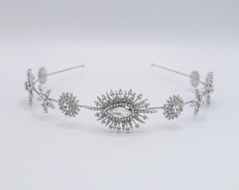 May include: A silver-toned celestial-themed headband. The design features a central oval-shaped crystal surrounded by radiating crystal accents, flanked by smaller star and floral-shaped embellishments. The headband is designed for formal occasions.