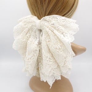 May include: A white lace hair bow with a large, layered bow. The bow is tied with a white ribbon and has a delicate, intricate lace pattern.