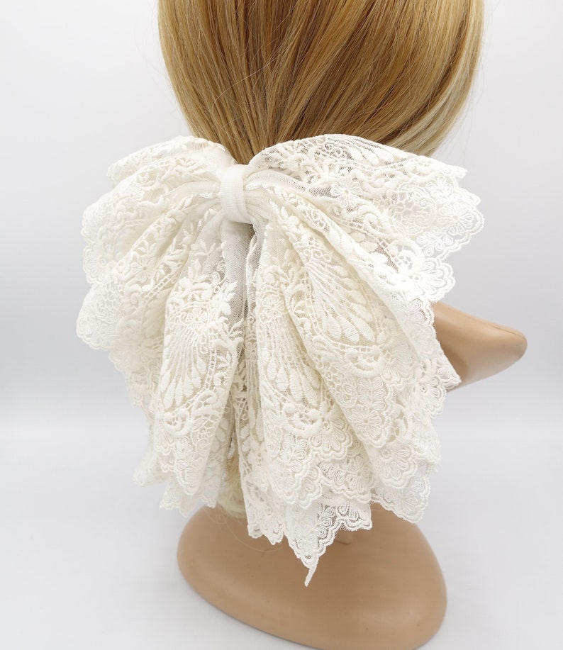 May include: A white lace hair bow with a large, layered bow. The bow is tied with a white ribbon and has a delicate, intricate lace pattern.