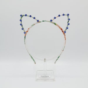 May include: A floral patterned headband with blue rhinestone cat ears. The headband is on a clear display stand with the brand name "VeryShine" printed on the base.