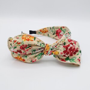May include: A beige headband with a large floral print bow. The bow is tied in a knot and the headband is made of a soft fabric.