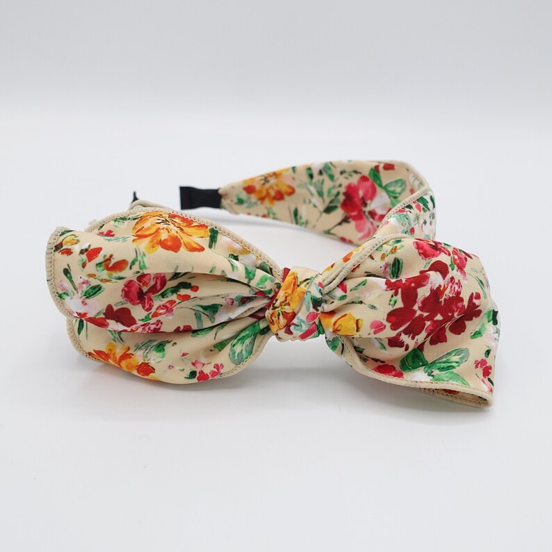 May include: A beige headband with a large floral print bow. The bow is tied in a knot and the headband is made of a soft fabric.