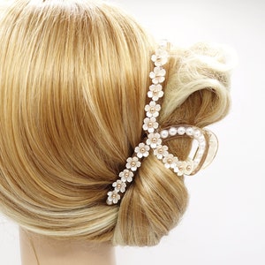 Puede incluir: Una pinza decorativa para el cabello con un diseño floral. La pinza está adornada con pequeñas flores blancas y detalles dorados. La pinza sujeta un moño de pelo rubio.