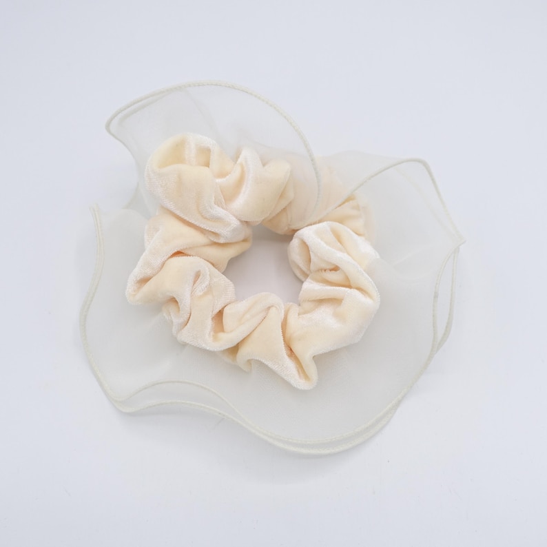 May include: A cream-colored scrunchie made of soft velvet fabric, centered on a white, ruffled organza base. The scrunchie is a hair accessory, perfect for adding a touch of elegance to any hairstyle.