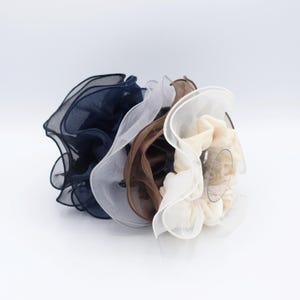 May include: A collection of four scrunchies in various colors: navy blue, white, brown, and cream. Each scrunchie is made of a sheer, ruffled fabric, creating a voluminous and textured appearance. The scrunchies are arranged in a cluster, showcasing their different hues.