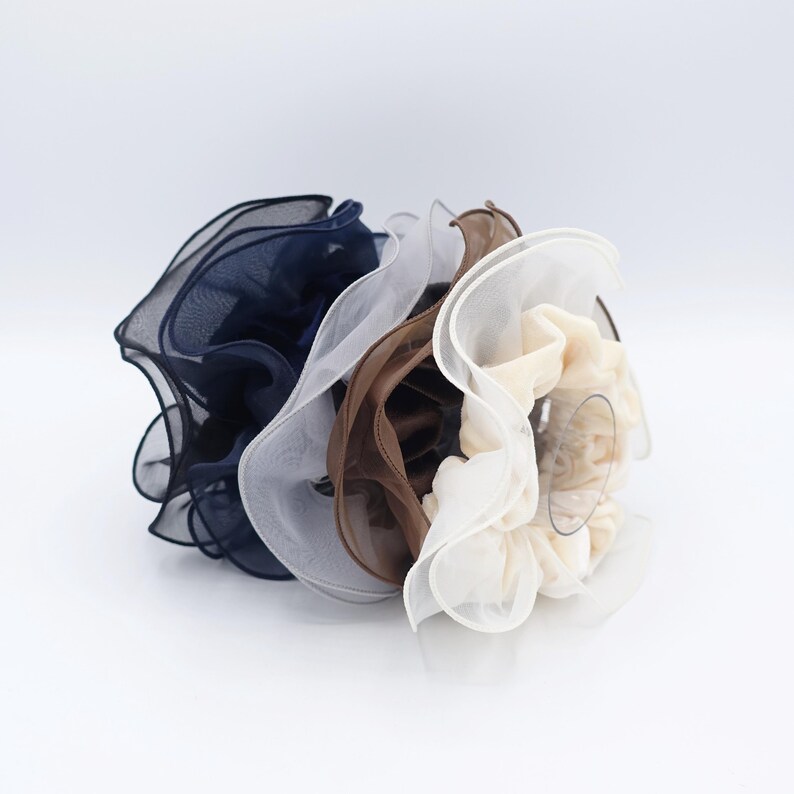 May include: A collection of four scrunchies in various colors: navy blue, white, brown, and cream. Each scrunchie is made of a sheer, ruffled fabric, creating a voluminous and textured appearance. The scrunchies are arranged in a cluster, showcasing their different hues.