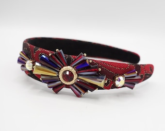 May include: Red headband with a large, gold-toned, jewel-encrusted flower design. The flower has a red center stone and is surrounded by iridescent, multi-colored crystals.