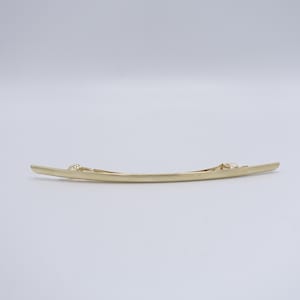 May include: A long, slender, gold-toned metal hair barrette with a smooth, slightly curved design. The accessory features a simple, polished finish and a functional spring-loaded clip mechanism attached to the back for securing hair in place.