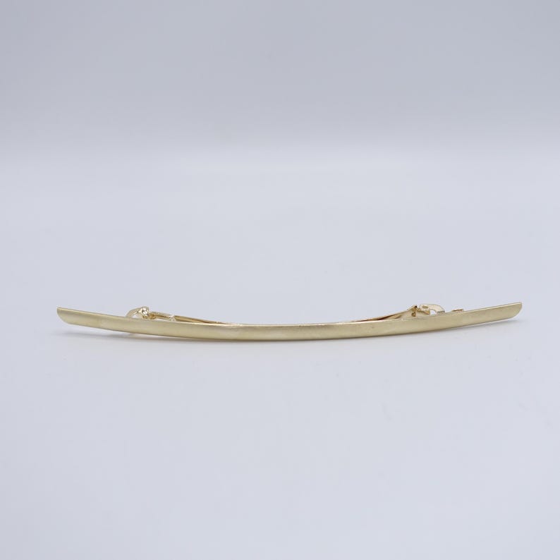 May include: A long, slender, gold-toned metal hair barrette with a smooth, slightly curved design. The accessory features a simple, polished finish and a functional spring-loaded clip mechanism attached to the back for securing hair in place.