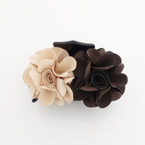 May include: A hair claw clip with two flower-shaped clusters. One cluster is beige, the other is dark brown. The clip is black. The flowers have a layered petal design, creating a textured appearance.