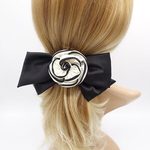 May include: A black satin bow with a white and black striped flower accent. The bow is attached to a hair clip.