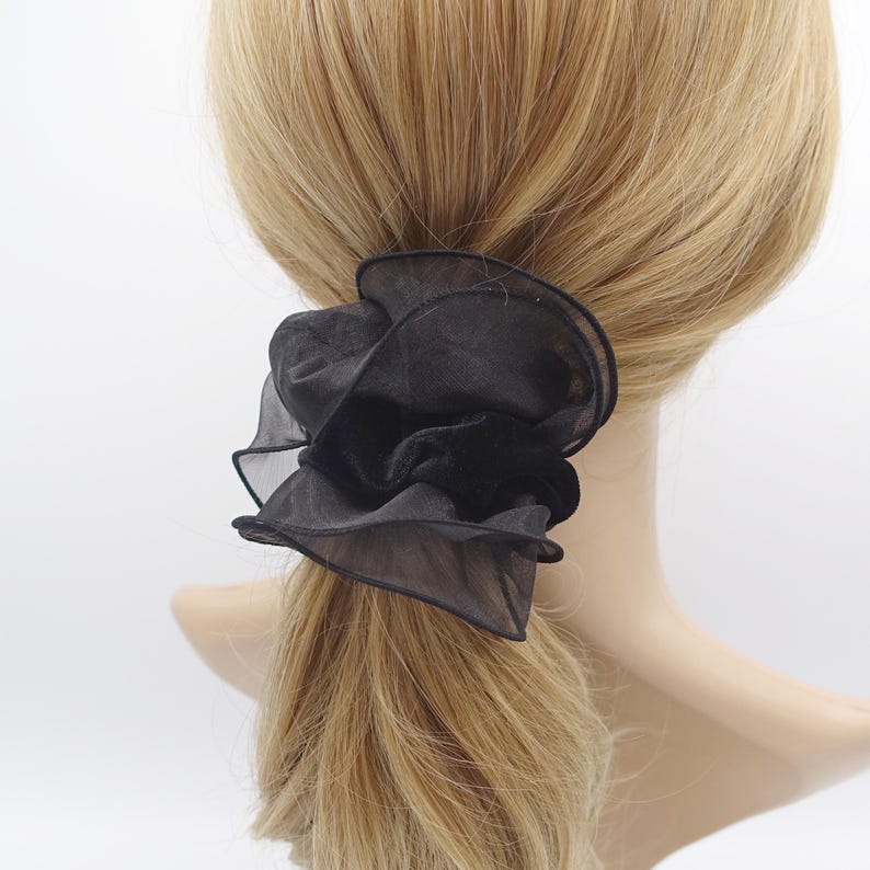 May include: A black scrunchie with a layered, ruffled design. The scrunchie is made of a sheer fabric, creating a voluminous, flower-like appearance. The hair accessory is attached to blonde hair.