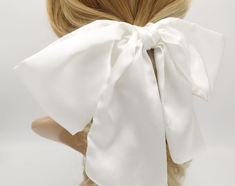 Op de afbeelding: Een witte satijnen haarband met een grote, vloeiende strik. De strik is om een ​​paardenstaart gebonden en is een eenvoudig, elegant haaraccessoire.
