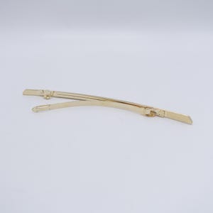 May include: A gold-toned metal hair barrette featuring a slender, curved design. The accessory consists of a smooth, arched top piece connected to a hinged clasp mechanism that secures the hair in place with a simple, functional metal closure.
