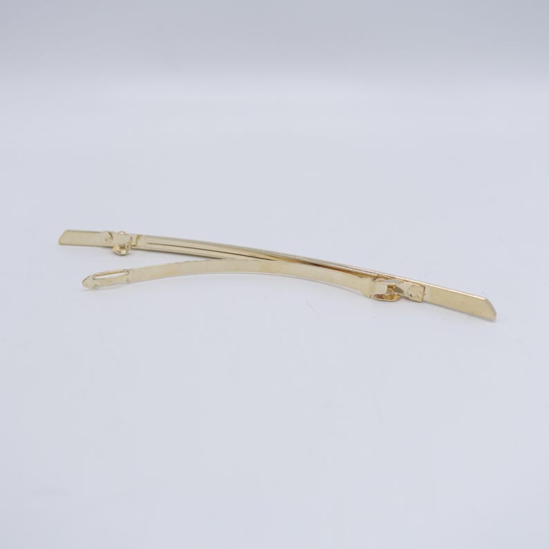 May include: A gold-toned metal hair barrette featuring a slender, curved design. The accessory consists of a smooth, arched top piece connected to a hinged clasp mechanism that secures the hair in place with a simple, functional metal closure.