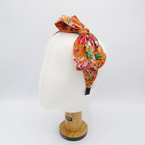 May include: Orange headband with a large floral print bow. The bow is made of fabric and has a ruffled edge. The headband is black.