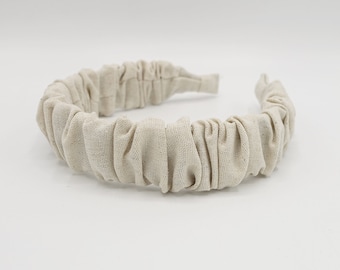 May include: A beige fabric headband with a gathered or ruffled design. The headband is made of a soft, textured fabric.
