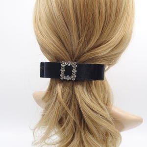 Puede incluir: Un lazo para el cabello de terciopelo negro con una hebilla rectangular adornada con pedrería. El accesorio para el cabello está sujeto a cabello rubio, lo que sugiere un estilo formal. Un accesorio clásico.
