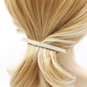 May include: A gold hair clip with clear rhinestones. The clip is in the shape of a long rectangle and is being worn in a woman's blonde hair.